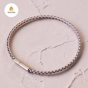 Op de afbeelding: Een gevlochten armband met een lichtpaars en goudkleurig ontwerp. De armband heeft een magnetische sluiting en is cirkelvormig. De armband ligt op een getextureerd, gebroken wit oppervlak.