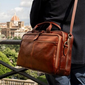 May include: A brown leather briefcase featuring a rectangular shape, dual top handles, and a detachable shoulder strap with metal hardware. The bag includes a front zippered pocket and multiple compartments with visible stitching along the edges.