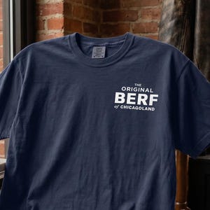 May include: A navy blue short-sleeve t-shirt featuring white text on the upper left chest that reads THE ORIGINAL BERF of CHICAGOLAND. The garment has a crew neckline and a standard fit, displayed on a wooden chair in a city setting.