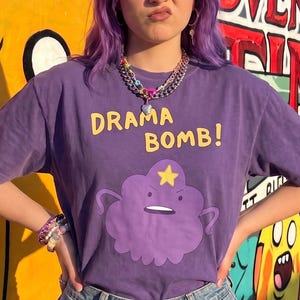 May include: A purple short-sleeved t-shirt featuring a graphic of a purple cloud character with a yellow star on its head. The text DRAMA BOMB! is printed in yellow above the character. The shirt is paired with light blue denim shorts.
