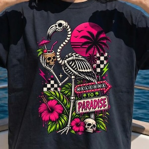 May include: A black t-shirt featuring a graphic print of a skeleton flamingo holding a coconut drink, surrounded by palm trees, hibiscus flowers, and a skull. A sign reads "WELCOME TO PARADISE" in pink and white text against a vibrant sunset background.