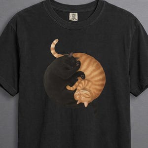May include: A black short-sleeved t-shirt featuring a graphic print of a solid black cat and an orange tabby cat curled together in a circular yin-yang formation. The cats are positioned in the centre of the chest on the dark fabric.