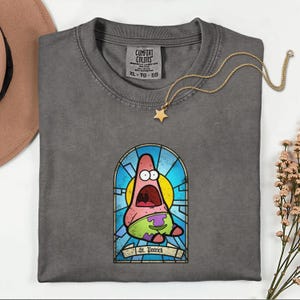 Peut inclure: Un t-shirt gris Comfort Colors avec un motif de vitrail de Patrick Star de Bob l'éponge. Le motif comprend le texte "St. Patrick". Un collier pendentif étoile en or est posé sur le t-shirt.