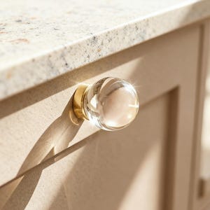 May include: A clear, round glass cabinet knob with a gold-colored base. The knob is attached to a light beige cabinet door. The countertop above the cabinet is a speckled beige and white granite.