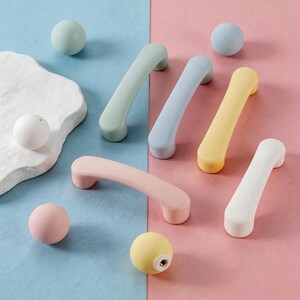 May include: A selection of pastel-coloured cabinet handles and spherical knobs in shades of green, blue, yellow, pink, and white. The hardware is arranged on a two-tone blue and pink background, creating a visually appealing display.