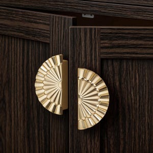 May include: Close-up of two gold-colored, fan-shaped cabinet pulls. The pulls are mounted on a dark brown wooden cabinet door. The pulls have a detailed, textured design, and the wood grain is visible.
