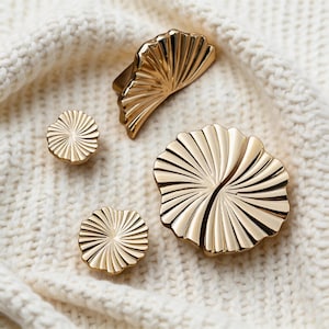 Może przedstawiać: Zestaw złotych elementów dekoracyjnych, w tym gałki do szafek i uchwyt do szuflady, ułożonych na kremowej dzianinie. Okucia mają wachlarzowaty kształt z promienistymi liniami, dodając elegancji.