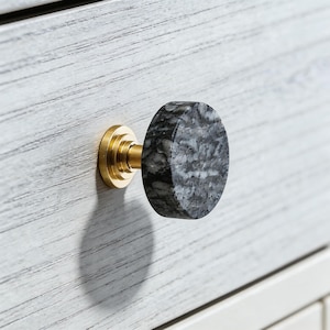 May include: A cabinet knob with a gray and white marble face and a gold-colored base. The knob is mounted on a white wooden cabinet with a horizontal grain pattern. The knob is round and has a geometric shape.