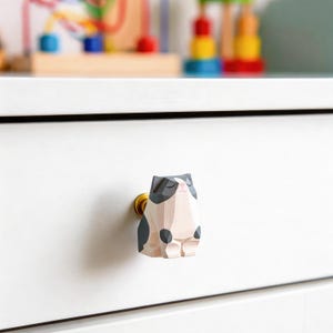 以下が含まれることがあります： 金色のベースが付いた、猫の形をした木製の引き出しつまみ。猫はグレー、白、ピンクの色合いで塗装されており、目は閉じられています。引き出しつまみは白い引き出しに取り付けられています。全体的なテーマは、気まぐれな動物をテーマにした家の装飾アイテムです。