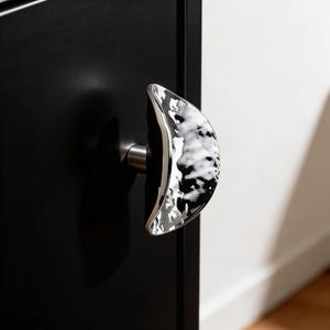 May include: A contemporary door handle with a distinctive design. The handle has a polished silver finish and a marbled black and white accent. It's mounted on a black door, creating a contrast. The handle's shape is curved and ergonomic, with a length of approximately 15 cm.