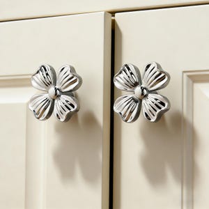 Op de afbeelding: Twee zilverkleurige, bloemvormige kastknoppen zijn gemonteerd op crèmekleurige kastdeuren. Elke knop heeft vier bloemblaadjes en een centraal, afgerond detail. De knoppen zijn reflecterend, wat wijst op een gepolijste metalen afwerking.