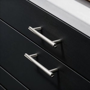May include: Close-up of two brushed nickel cabinet pulls on a black cabinet. The pulls are horizontal, cylindrical, and have a simple, modern design. The cabinet has a dark, textured finish.
