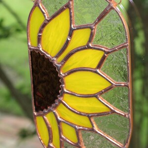 May include: An oval-shaped stained glass suncatcher depicting a sunflower. The petals are yellow, the centre is dark brown, and the glass is framed with copper-coloured metal. It hangs from a chain.