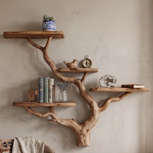 Op de afbeelding: Een uniek, boomvormig wandrek van hout met meerdere plateaus. De planken bevatten decoratieve items zoals een kleine potplant, boeken, een houten vogel, een vintage klok, een geode en een klein boek. Het rek is aan een neutraal gekleurde muur bevestigd.
