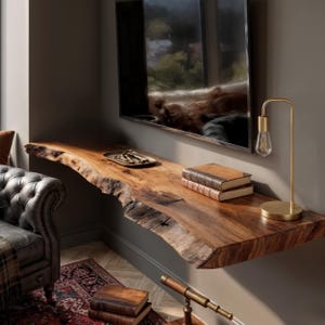 Puede incluir: Una estantería flotante de madera con borde natural e irregular está montada en una pared gris. Un televisor de pantalla plana está montado encima de la estantería. Una lámpara dorada y dos libros están sobre la estantería. También hay una pequeña bandeja con llaves.