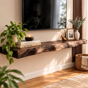 Puede incluir: Un estante flotante largo de madera de color marrón oscuro con borde natural montado en la pared. El estante sostiene una planta verde en maceta, una vela negra, dos libros apilados titulados The Kinfolk Home y At Home, un pájaro de madera y un jarrón.