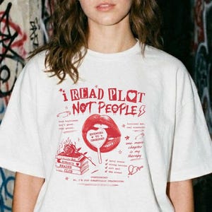 May include: A white short-sleeve t-shirt featuring a red graphic print that reads I READ PLOT NOT PEOPLE. The design includes illustrations of a pair of lips, a stack of books, and various small symbols like a safety pin, a heart, and a lightning bolt.