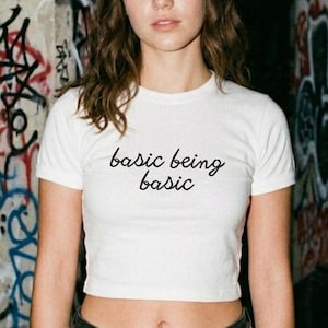 May include: A white short-sleeved crop top t-shirt featuring the black cursive text basic being basic printed on the center chest. The garment has a crew neckline and a fitted silhouette, paired with dark denim shorts featuring a silver chain belt.