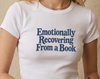 May include: A white short-sleeved ribbed t-shirt featuring the dark blue text "Emotionally Recovering From a Book" printed across the chest. The garment has a crew neckline and a fitted silhouette, showcasing a simple and clean design.