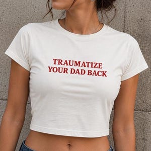May include: A white short-sleeved crop top featuring the red text "TRAUMATIZE YOUR DAD BACK" printed across the chest. The garment has a crew neckline and a fitted silhouette, paired with blue denim jeans and dark oval-shaped sunglasses.