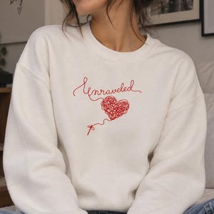 May include: A white crewneck sweatshirt featuring red embroidery on the chest. The design consists of the word Unraveled in cursive script above a heart shape formed by a tangled red thread, with a small needle attached to the end of the string.