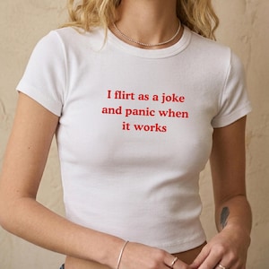 May include: White ribbed crop top with short sleeves. The text "I flirt as a joke and panic when it works" is printed in red. The top is a casual style.