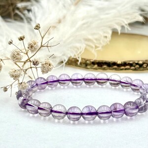 Puede incluir: Un brazalete de cuentas redondas de amatista translúcida. Las cuentas, de unos 0,8 cm de diámetro, son de color lila claro con rayas moradas más oscuras. El brazalete está sobre una superficie blanca, con elementos florales y dorados.