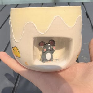 Peut inclure: Une tasse en céramique au design fantaisiste. La tasse présente un dessus jaune clair avec un effet dégoulinant, une petite figurine de souris à l'intérieur d'une découpe et une illustration de tranche de fromage. La tasse est tenue dans une main.