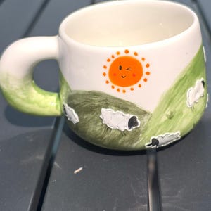 Pode incluir: Caneca de cerâmica branca com um design lúdico com um sol laranja sorridente, colinas verdes e várias ovelhas. A alça tem formato arredondado com detalhes verdes. Para bebidas quentes.