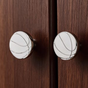 Op de afbeelding: Twee ronde kastknoppen met een basketbalontwerp. De knoppen hebben een zilveren afwerking en een wit oppervlak met zwarte lijnen die het patroon van een basketbal nabootsen. Gemonteerd op een donkerbruine houten kast.