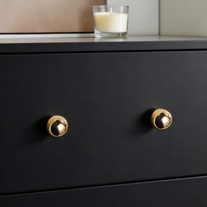 May include: A black dresser with four gold-colored, round knobs. The knobs are centered on the drawers. A lit candle in a glass holder sits on top of the dresser.