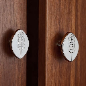 May include: Two cabinet knobs with a football-shaped design. The knobs have a white centre with a silver outline and a silver stem. The knobs are mounted on a dark brown wooden cabinet.