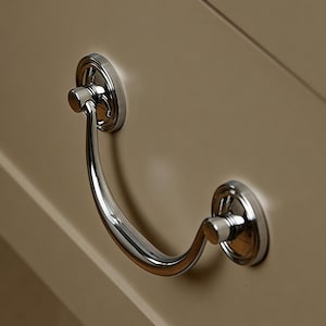 Könnte beinhalten: Nahaufnahme einer Schublade mit einem polierten Chromgriff. Der Griff ist gebogen und mit zwei kreisförmigen, dekorativen Halterungen an der Schublade befestigt. Die Schublade ist in einem neutralen Beige gestrichen.