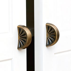 Puede incluir: Dos tiradores de armario de latón antiguo, cada uno con forma de semicírculo con un diseño de sol. Los tiradores están montados en una puerta de armario blanca, ofreciendo una estética vintage.