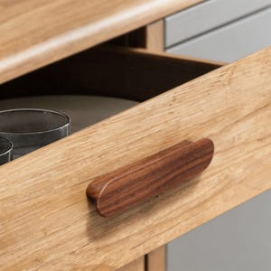 Op de afbeelding: Close-up van een open houten lade met een donkerbruine houten handgreep. De lade is gemaakt van lichtgekleurd hout en de handgreep heeft een contrasterende donkerdere tint. De lade lijkt deel uit te maken van een groter meubelstuk.
