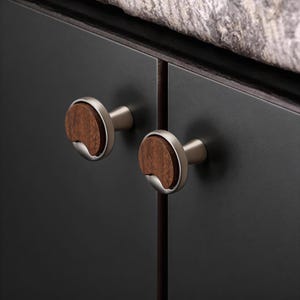 May include: Two cabinet knobs with a silver finish and a wooden, crescent-shaped center. The knobs are mounted on a dark gray cabinet door. The wood has a warm brown tone, and the silver has a matte finish.