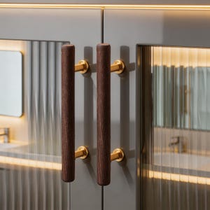 Puede incluir: Primer plano de un armario gris con dos tiradores de madera marrón oscuro y herrajes dorados. Las puertas del armario tienen una textura acanalada y están iluminadas con una luz cálida. El diseño sugiere un baño o una cocina modernos.