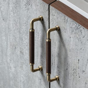 May include: Two cabinet handles with a vintage brass finish and dark brown leather-wrapped grips. The handles are mounted on a gray concrete-textured cabinet door with a dark wood trim.