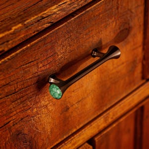 Pode incluir: Close-up de um puxador de gaveta em bronze escuro com um detalhe em pedra turquesa. O puxador está montado numa gaveta de madeira com grão visível e um tom castanho-avermelhado quente. O hardware tem um design moderno e elegante.
