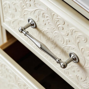 May include: Close-up of a cream-colored drawer with ornate floral carvings and a polished silver handle. The drawer is partially open, revealing the interior. The handle has a central, elongated section and rounded ends.