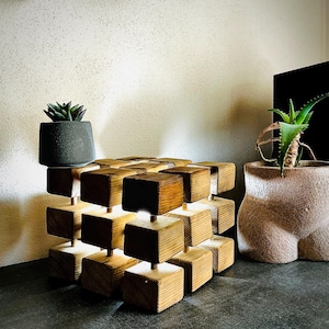 May include: A unique wooden cube lamp with a geometric design. The lamp is constructed from multiple wooden blocks, with warm light emanating from within. A small succulent plant in a dark gray pot sits atop the lamp. Another plant in a decorative pot is also visible.