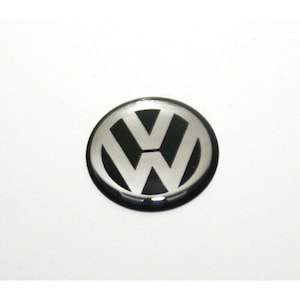 May include: A circular emblem featuring the Volkswagen logo in a metallic silver finish set against a black background. The design consists of a stylised V and W stacked vertically within a circular border, creating a high-contrast appearance.