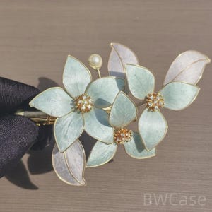 May include: A decorative hair clip with a floral design. The clip features light blue flower petals with gold detailing and a pearl. The flower centres are embellished with small crystals. The leaves are pale lavender.
