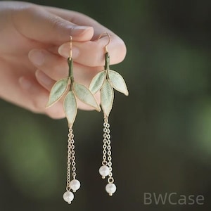 Op de afbeelding: Delicate oorbellen in de vorm van bladeren met een goudkleurige afwerking. De oorbellen hebben lichtgroene stoffen bladeren met gouden randen en bungelende kettingen met witte parelaccenten. Haaksluiting.