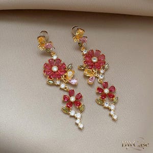 May include: A pair of gold-toned floral drop earrings. Each earring features a large, red flower with a central gem, smaller red flowers, and accents of white, yellow, and pink gems. The earrings have a delicate, elegant design.