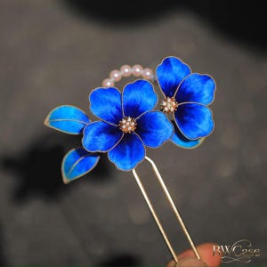 May include: A blue floral hair pin with gold detailing. The pin features two bright blue flowers with gold edging and a string of small pearls. The flower centres have a sparkling, gem-like detail. A decorative hair accessory.