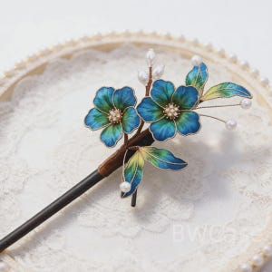 Könnte beinhalten: Dekorative Haarspange mit blauen und grünen Blumen und Perlenakzenten. Die Blumen haben goldene Zentren und sind an einem dunkelbraunen Stiel und einem schwarzen Stab befestigt. Ein Haarschmuck.