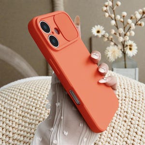 Puede incluir: Una funda de teléfono naranja con una cubierta para la cámara. La funda tiene un acabado liso y presenta recortes para las lentes de la cámara y los botones. La funda está diseñada para proteger el teléfono de arañazos e impactos.