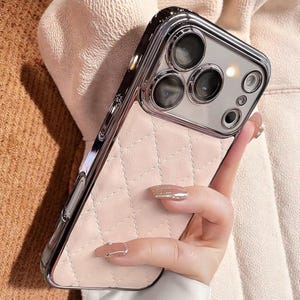 May include: A phone case with a quilted, light pink back and a shiny silver camera frame. The case has a diamond pattern and a metallic border. The phone case is held by a person.