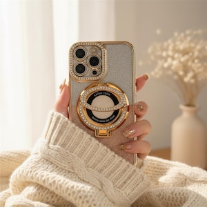 May include: A gold and silver glitter phone case with a ring holder. The case has a gold frame and is embellished with rhinestones around the camera and ring holder. The ring holder has the text "KATHY TECH" and "Quality makes the future".